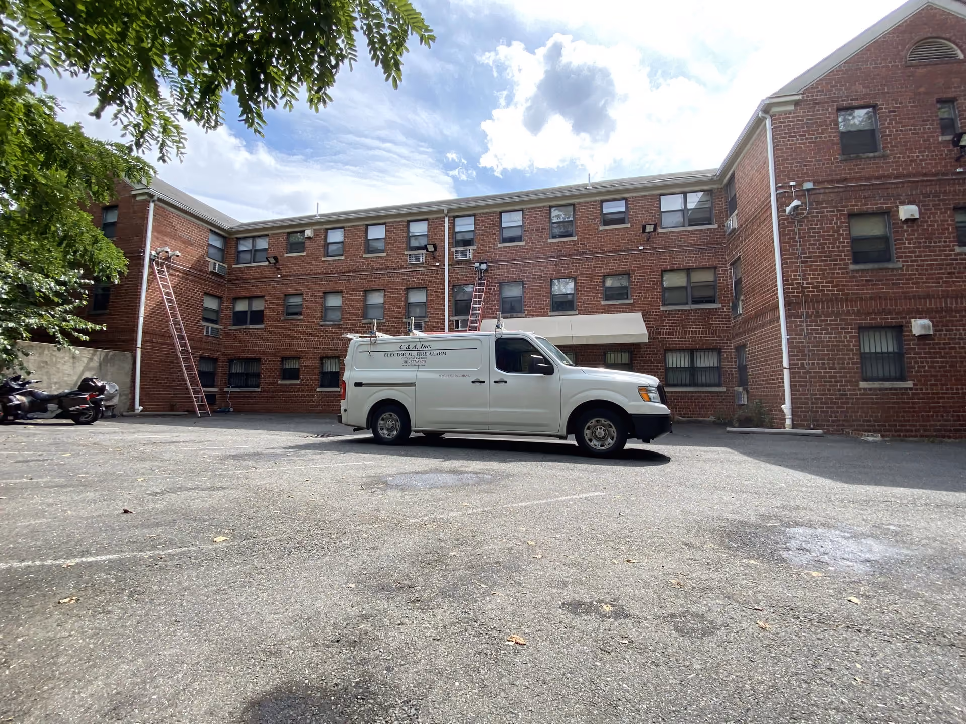 A C & A service van in the parking lot of a three-storey brick apartment building, with two extension ladders set against the wall.