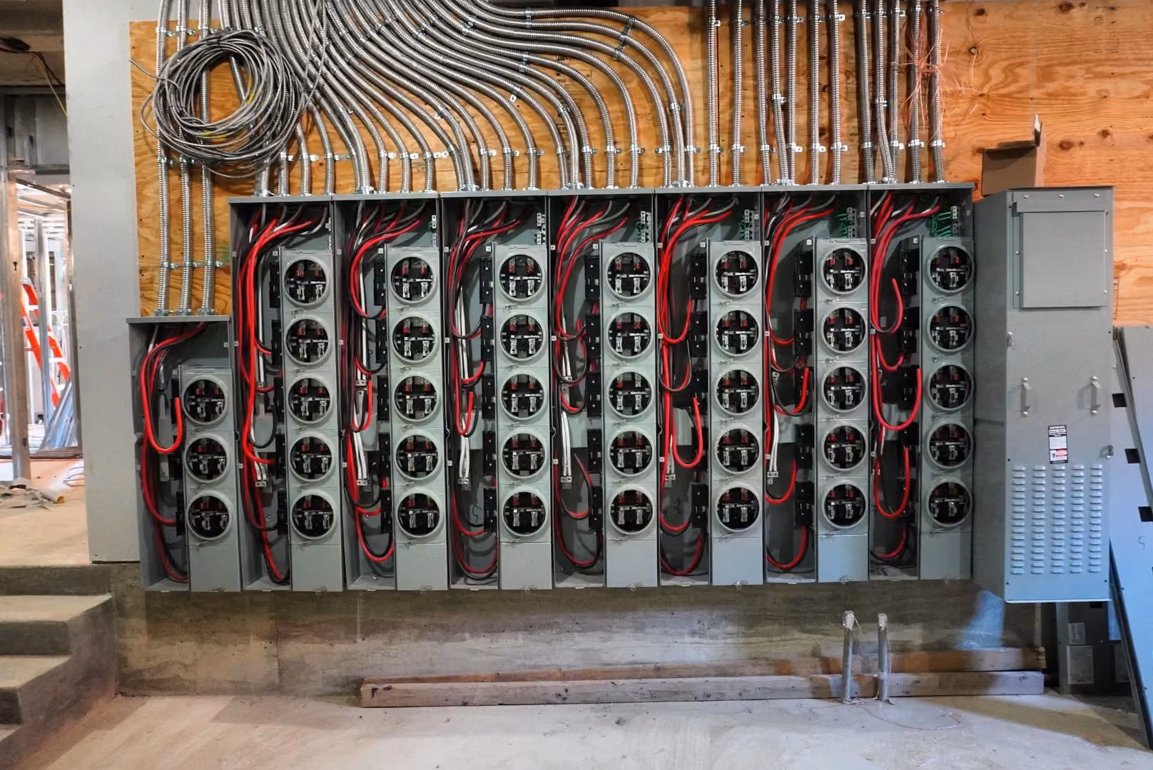 A bank of eight breaker panels wired with red conductors, fed by a fanned run of steel conduit across a plywood backboard.