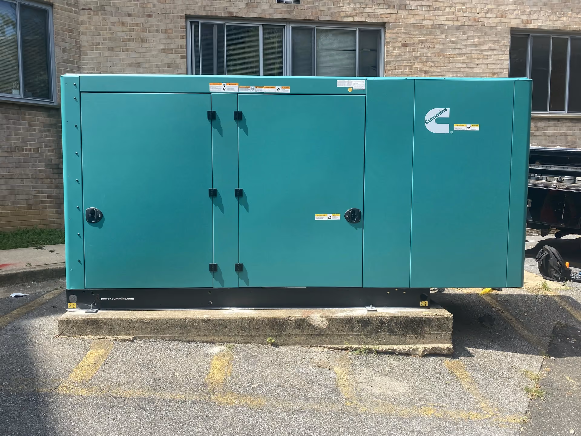 A Cummins standby generator in a green weatherproof enclosure, set on a concrete pad against a brick wall.
