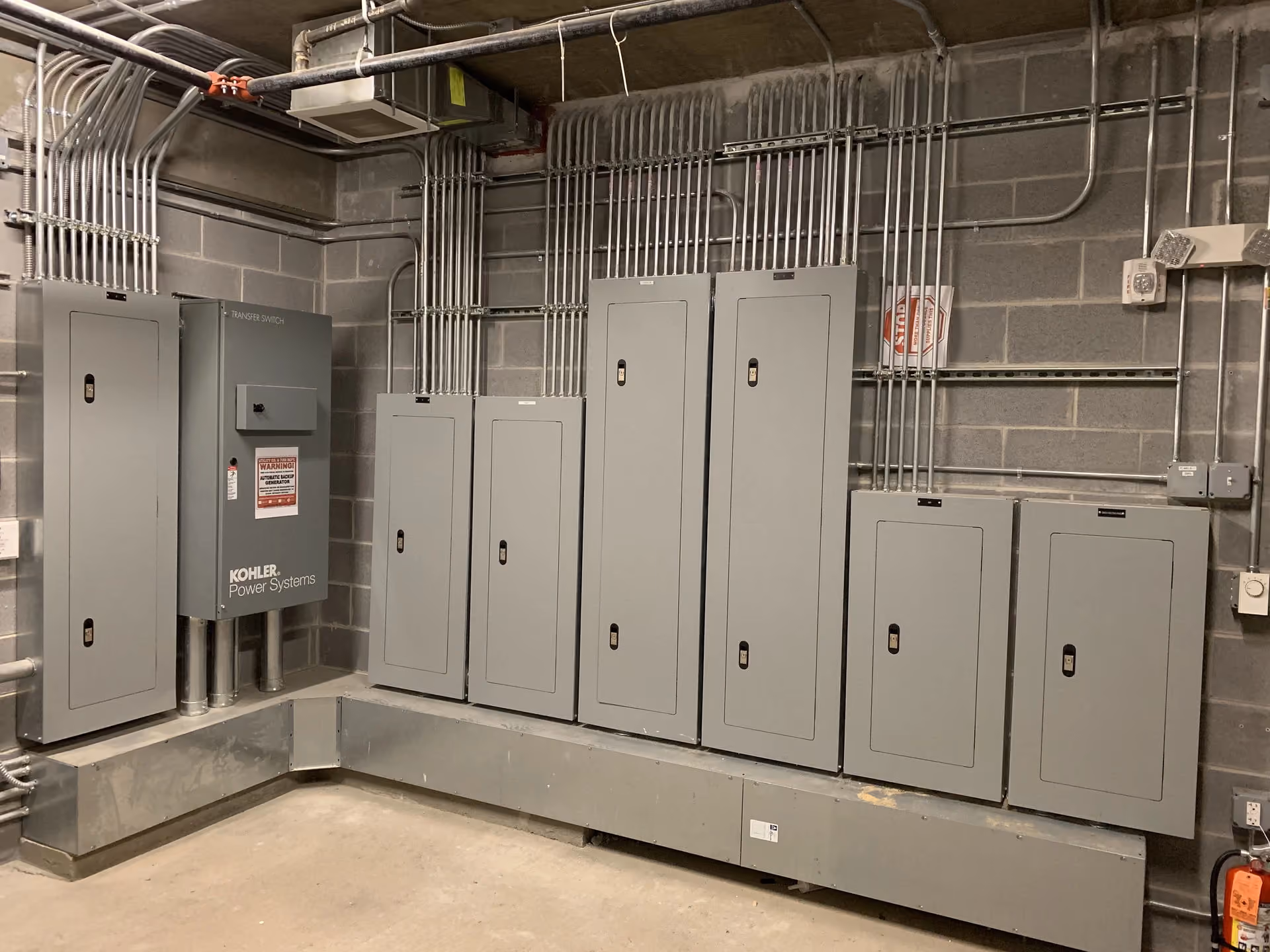 Kohler power-system switchgear beside a row of grey panelboards in a block-walled electrical room, with conduit banked overhead.