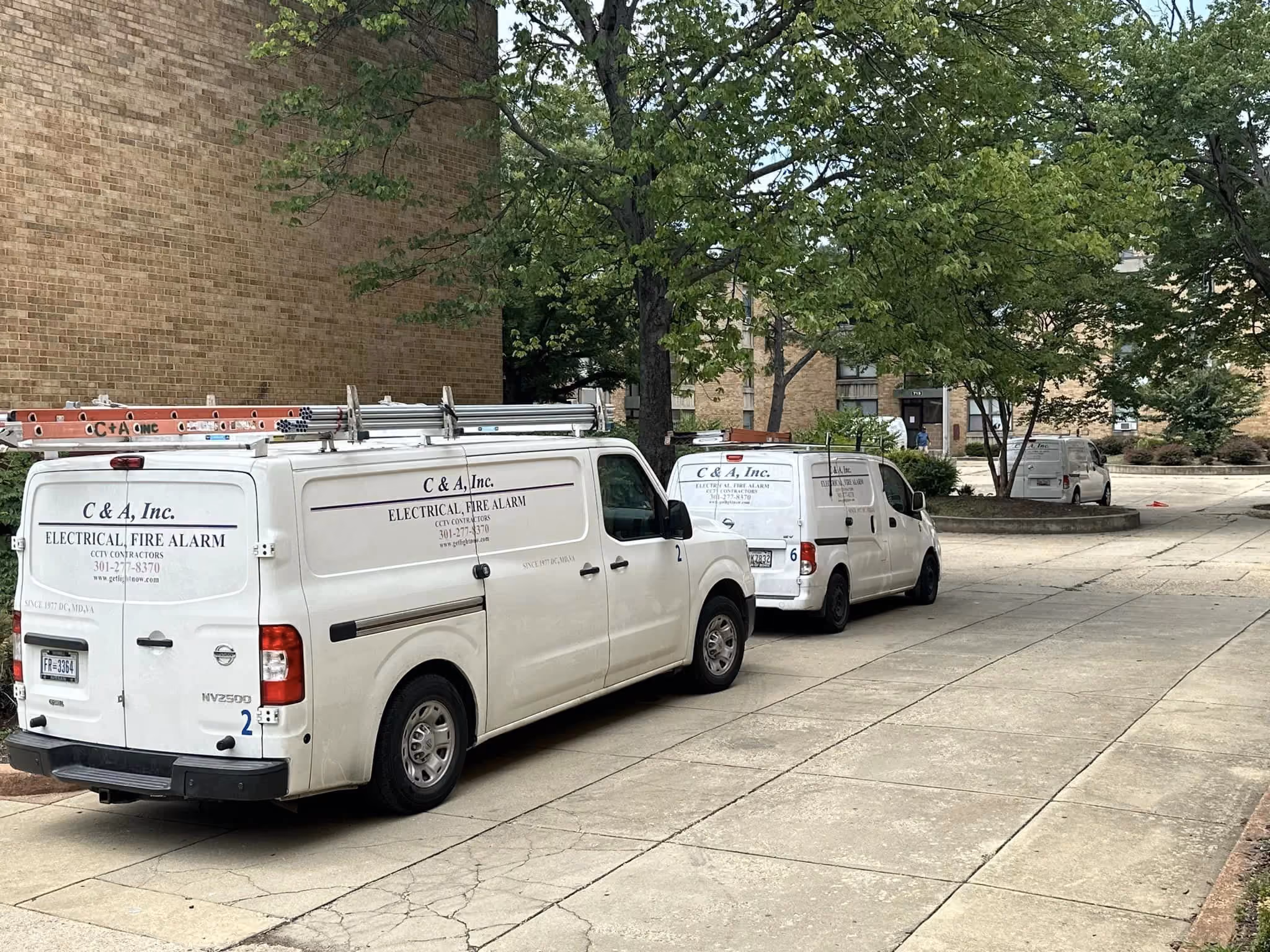 Two C & A service vans parked on a walkway beside a brick apartment building, ladders racked on the roof of each.